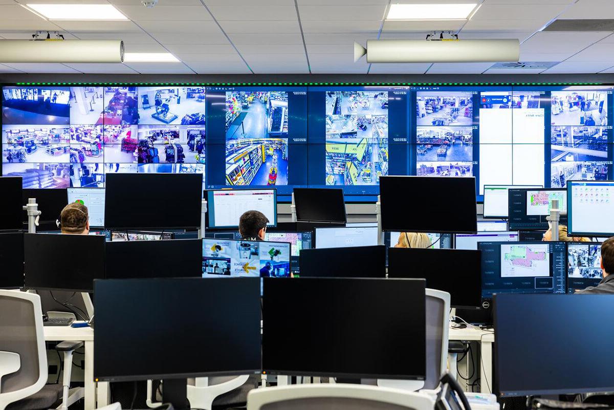 各种各样的人在办公桌前工作，面前是一面墙的电脑屏幕, showing a range of CCTV imagery